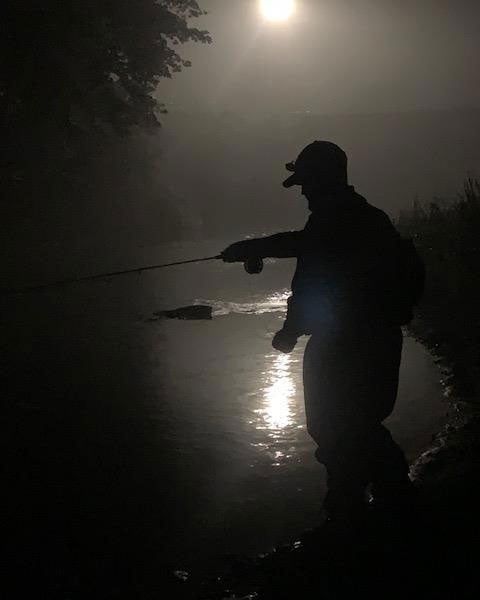 Silhouetted angler casting beside a moonlit river at night